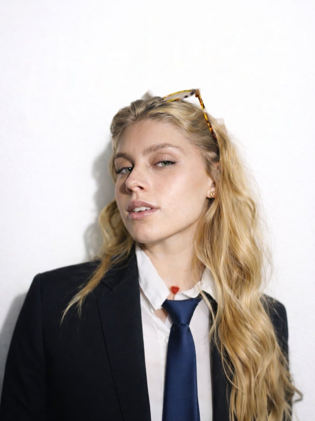 Ana Luiza, de blazer preto, camisa branca e gravata azul, olhando para a câmera