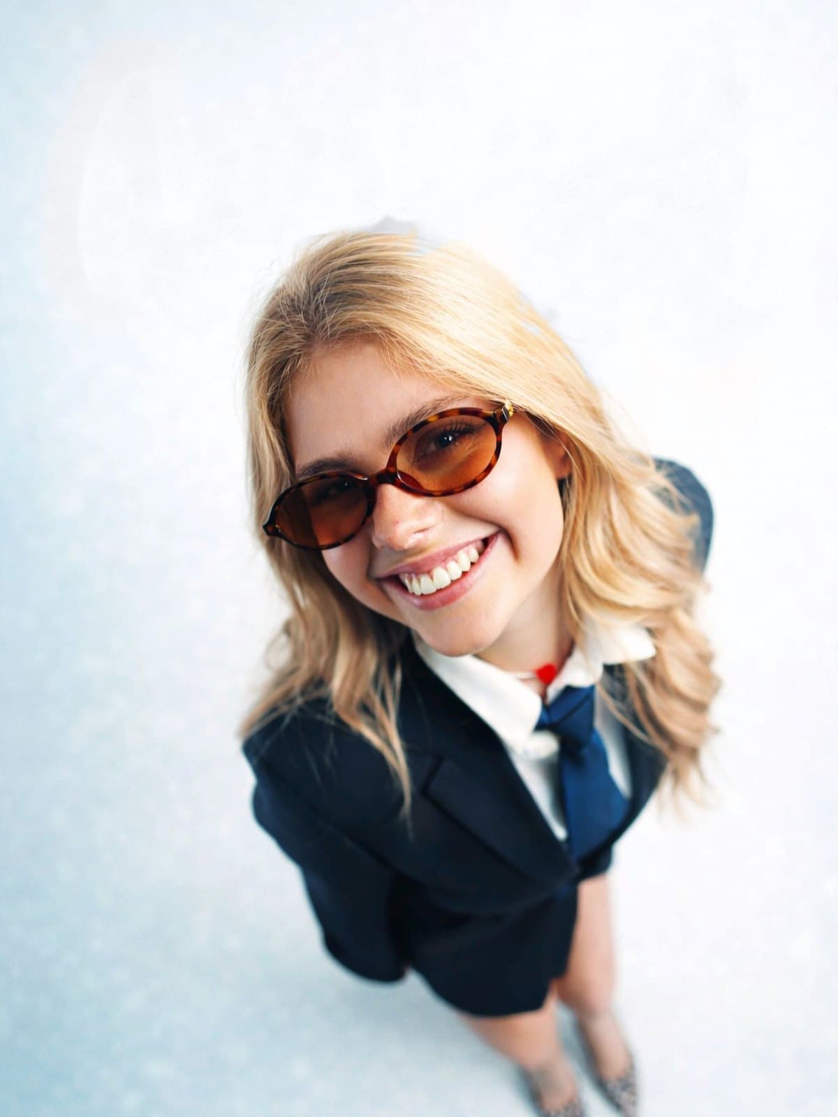 Ana Luiza sorrindo, fotografada de cima, de óculos escuros e blazer azul-marinho