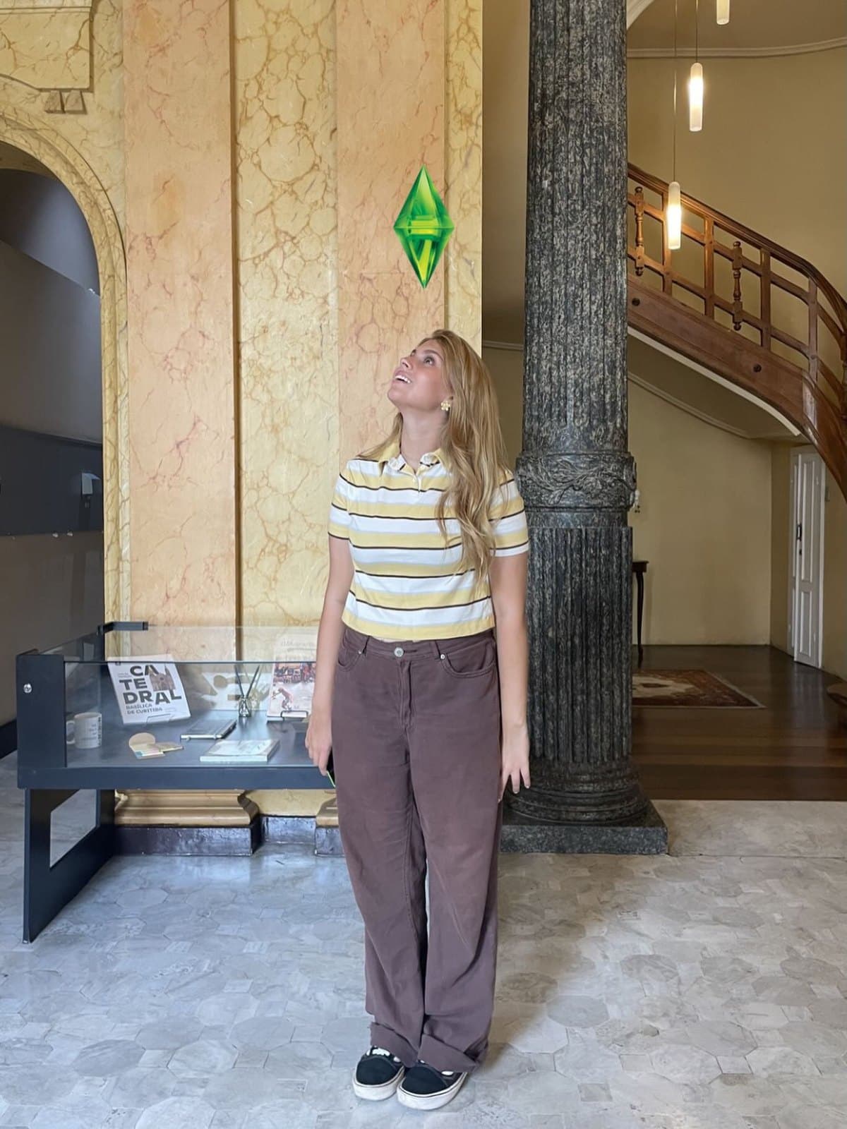 Ana Luiza em um saguão amplo, olhando para cima, com um cristal verde flutuando sobre a cabeça